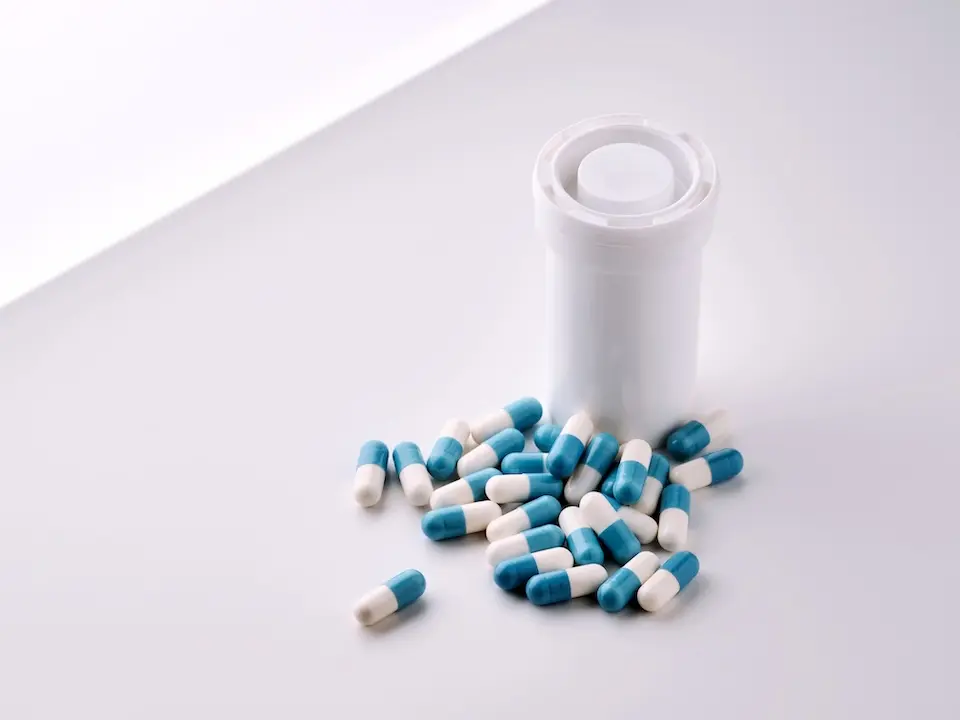 Frasco branco de medicamento ao lado de cápsulas azul e branca espalhadas sobre superfície clara