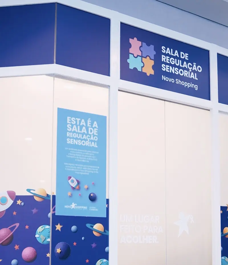 Fachada da Sala de Regulação Sensorial com letreiro azul 'Sala de Regulação Sensorial Novo Shopping' e adesivos com elementos espaciais nas portas de vidro