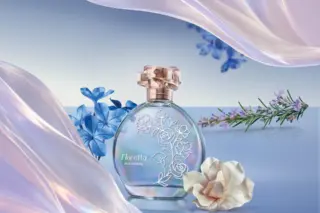 Frasco redondo do perfume Floratta Blue Crystal com acabamento iridescente, sobre superfície refletiva, cercado por flores azuis, uma flor branca e um ramo com pequenas flores lilás; tecido translúcido lilás em primeiro plano