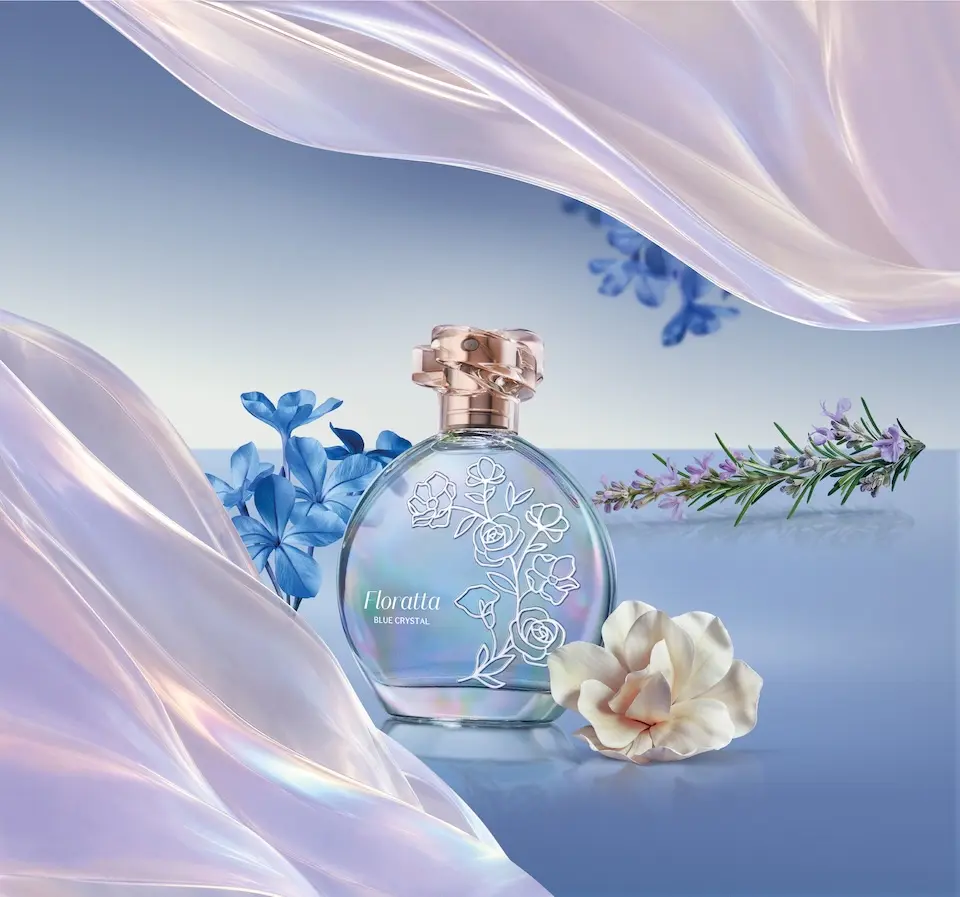 Frasco redondo do perfume Floratta Blue Crystal com acabamento iridescente, sobre superfície refletiva, cercado por flores azuis, uma flor branca e um ramo com pequenas flores lilás; tecido translúcido lilás em primeiro plano