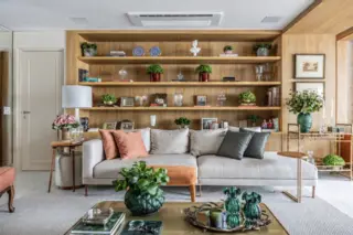 Sala de estar com sofá claro, almofadas em tons de laranja e verde, estante de madeira com objetos decorativos e mesa de centro com vasos verdes