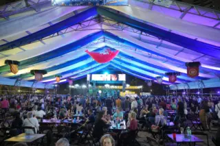 Tenda grande iluminada em tons de azul e roxo com público em mesas e pessoas em pé diante de um palco com painel de LED