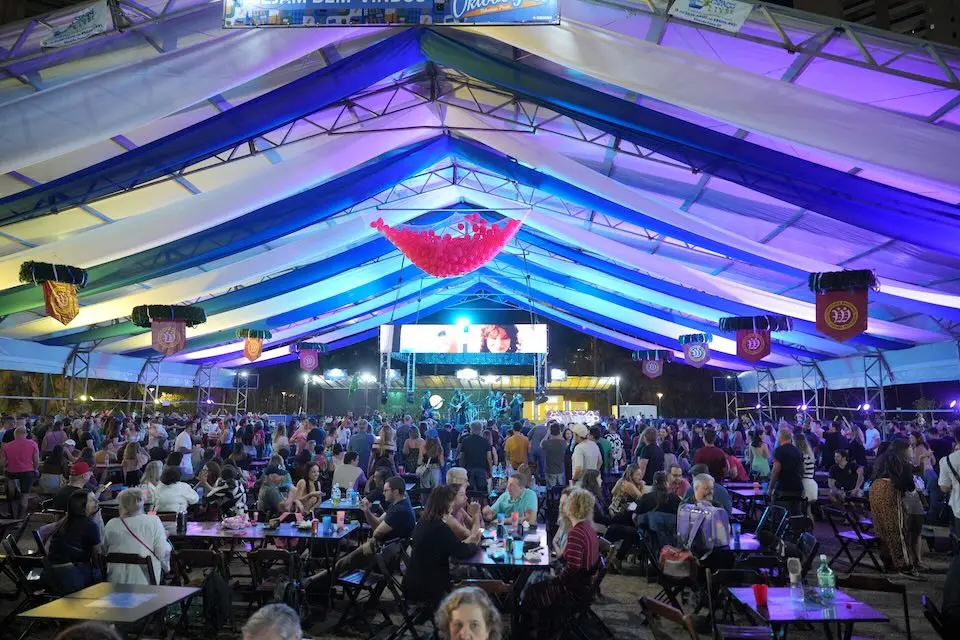 Tenda grande iluminada em tons de azul e roxo com público em mesas e pessoas em pé diante de um palco com painel de LED