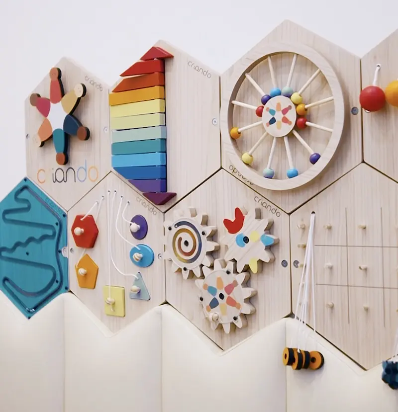 Painéis sensoriais Montessori em madeira com módulos hexagonais coloridos e elementos móveis, acima de painel estofado branco