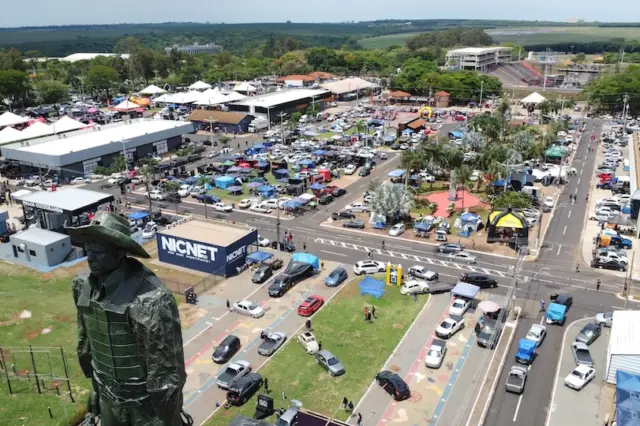 estátua de um peão em primeiro plano e vista aérea do Parque do Peão com veículos, tendas e estandes