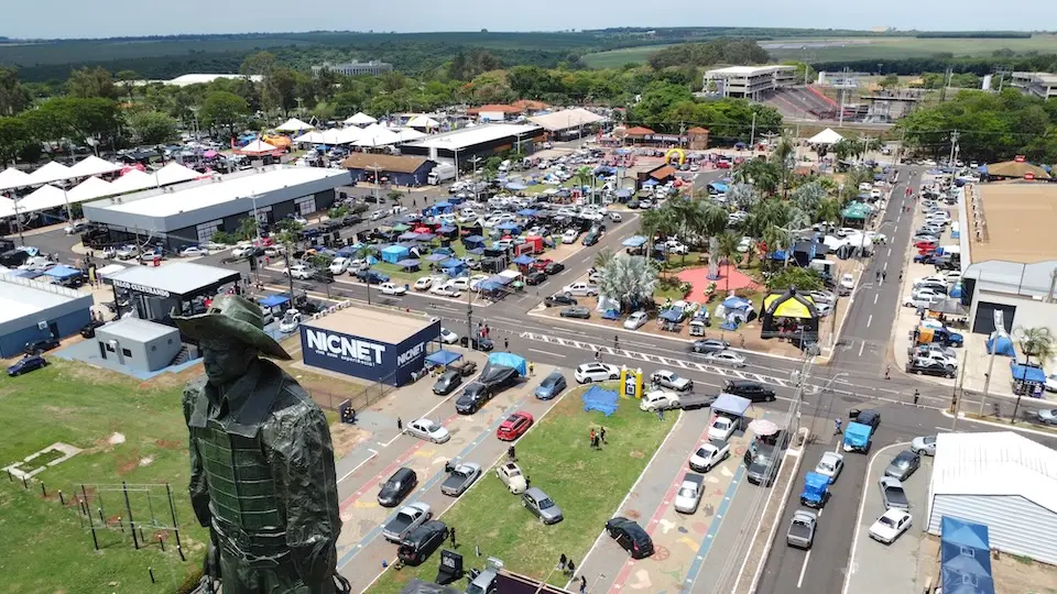estátua de um peão em primeiro plano e vista aérea do Parque do Peão com veículos, tendas e estandes