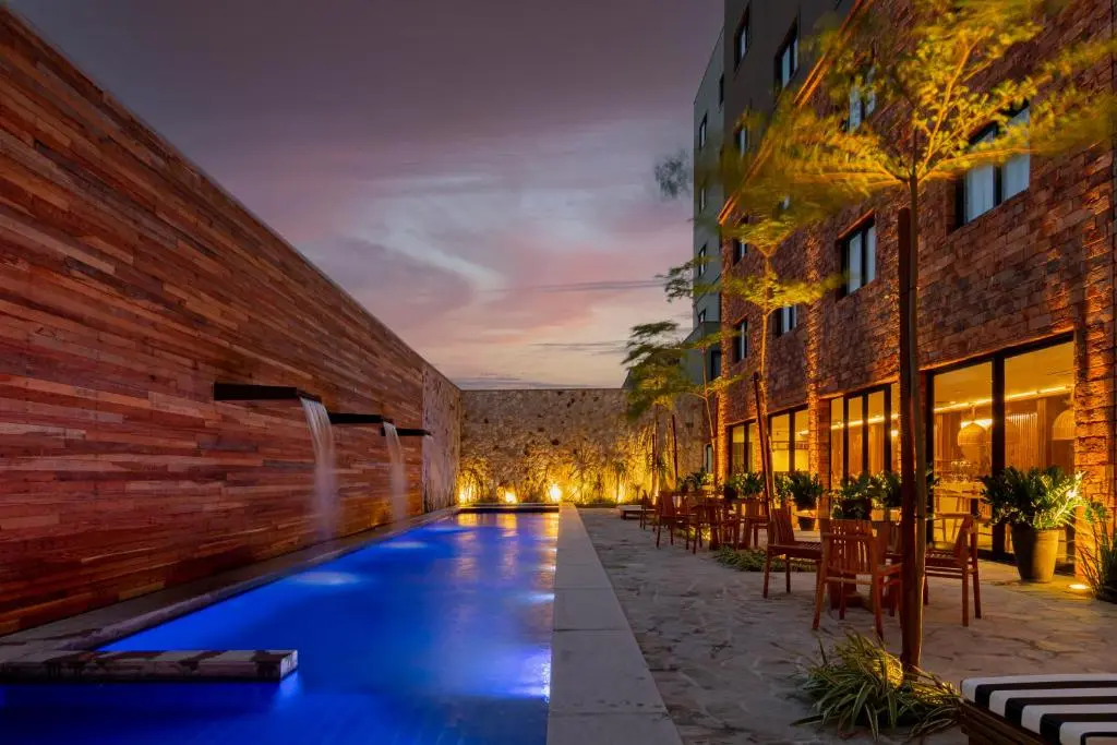 piscina externa iluminada em azul junto a parede de madeira com três quedas d'água, ao lado de fachada de hotel com janelas iluminadas