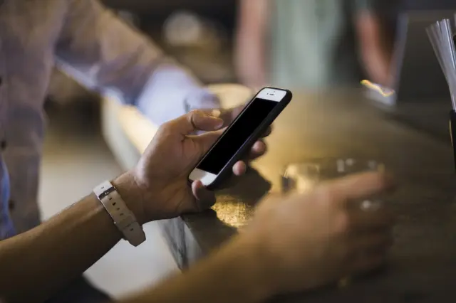 Mão segurando smartphone com tela apagada sobre balcão, com copo e outra mão desfocados ao fundo
