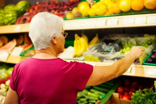 Mulher idosa vista de costas, com cabelo branco e blusa rosa, alcançando caixas de uvas na seção de hortifrúti de um supermercado