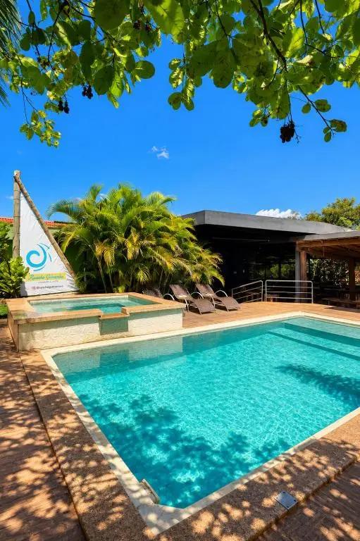 Piscina retangular com água azul, deck de madeira e espreguiçadeiras, com vegetação e vela triangular ao fundo