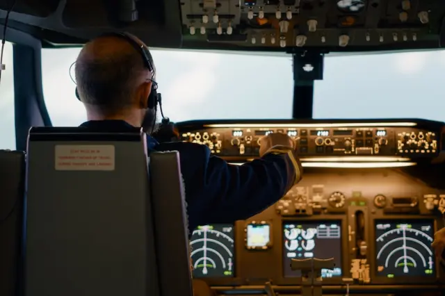Piloto com headset visto de costas no cockpit de uma aeronave, com painel de instrumentos iluminado