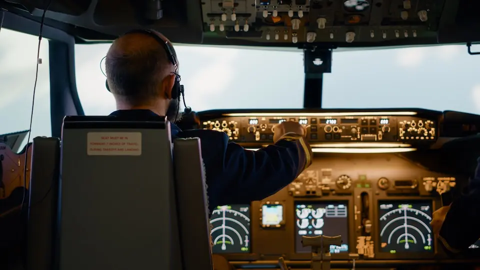 Piloto com headset visto de costas no cockpit de uma aeronave, com painel de instrumentos iluminado