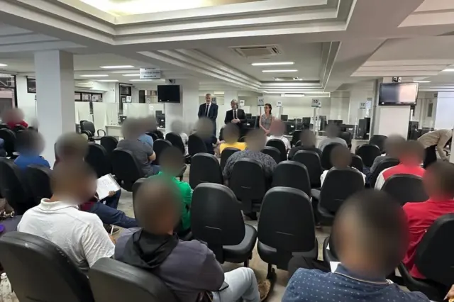 Pessoas sentadas de costas em fileiras de cadeiras em ampla sala de atendimento, com três pessoas em pé à frente