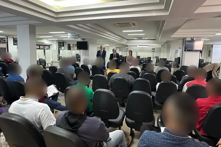 Pessoas sentadas de costas em fileiras de cadeiras em ampla sala de atendimento, com três pessoas em pé à frente