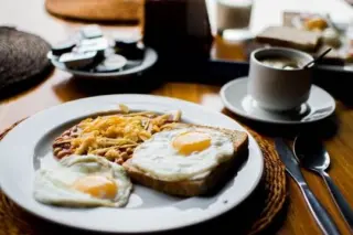 Prato com ovos fritos, torrada com ovo e porção de feijão ao lado de xícara de café e utensílios sobre mesa