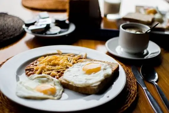 Prato com ovos fritos, torrada com ovo e porção de feijão ao lado de xícara de café e utensílios sobre mesa