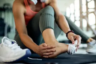 Pessoa sentada tirando a meia do pé direito, com tênis brancos ao lado e equipamentos de ginástica desfocados ao fundo