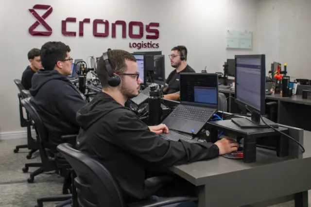 Pessoas sentadas em estações de trabalho com headsets e monitores; logo 'cronos logistics' em destaque na parede
