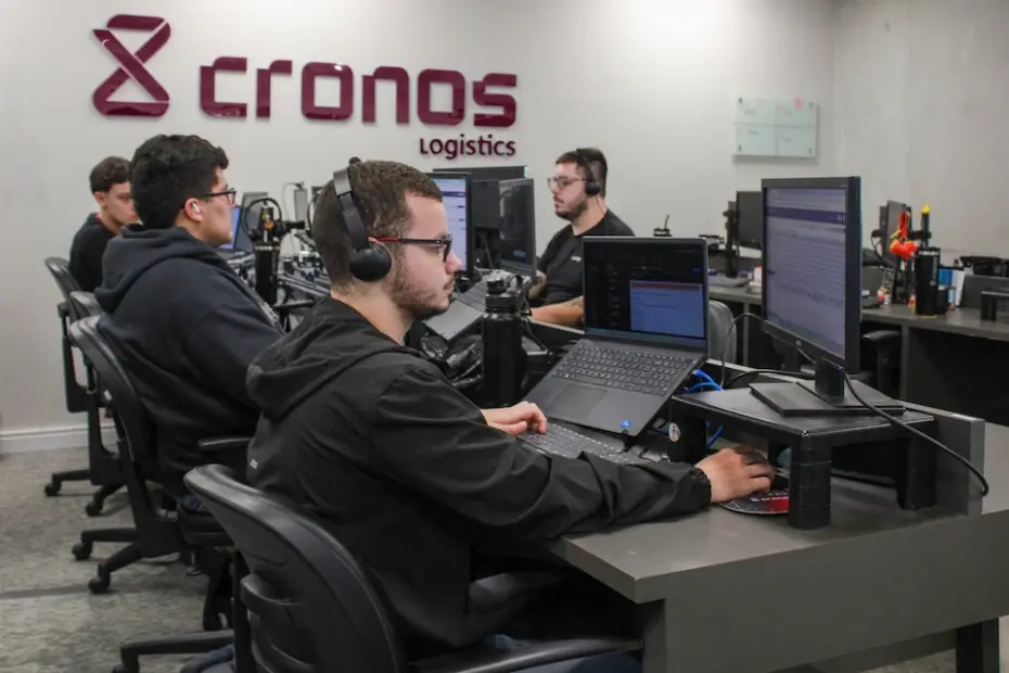 Pessoas sentadas em estações de trabalho com headsets e monitores; logo 'cronos logistics' em destaque na parede
