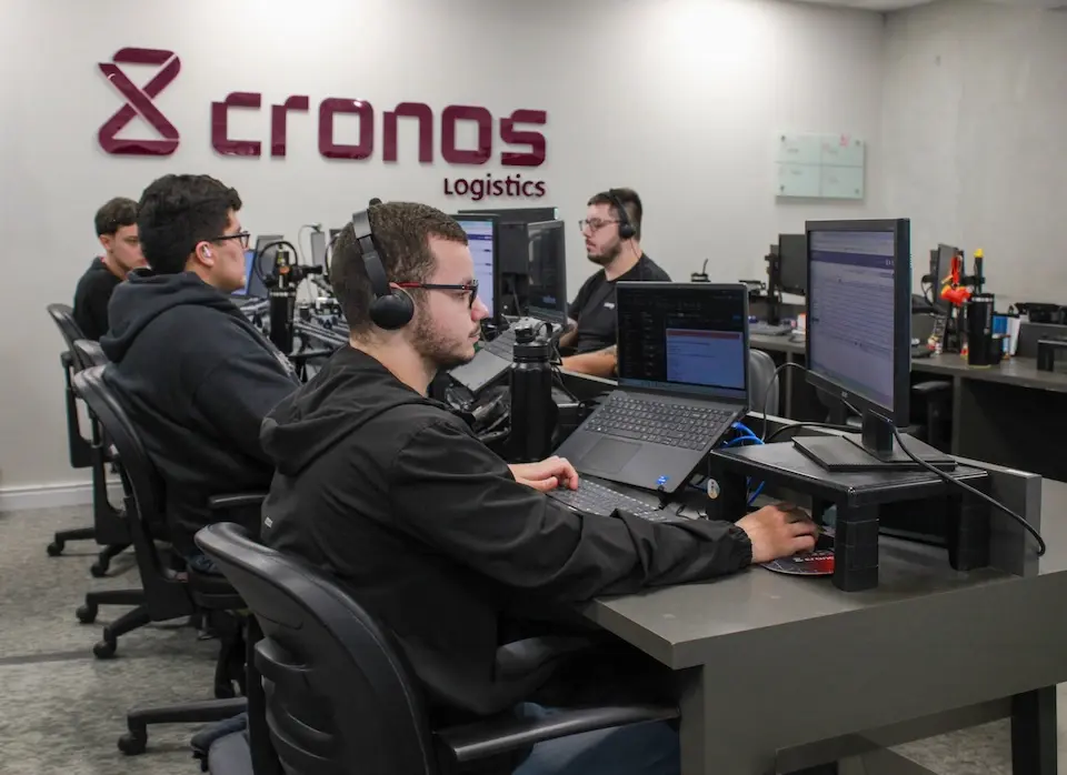 Pessoas sentadas em estações de trabalho com headsets e monitores; logo 'cronos logistics' em destaque na parede