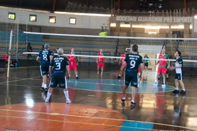 Partida de vôlei em quadra coberta com jogadores de duas equipes e bola no ar