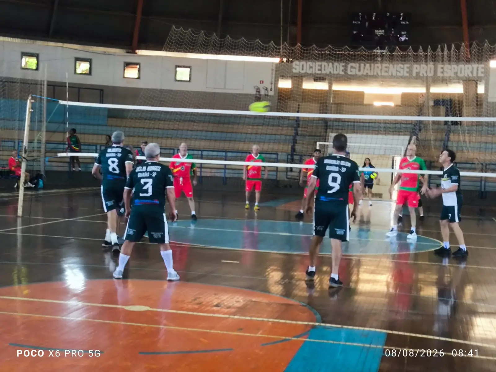 Partida de vôlei em quadra coberta com jogadores de duas equipes e bola no ar