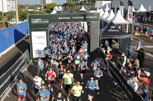 largada de corrida com corredores passando por arco de largada com as marcas Santander e Track&Field, tendas e público ao lado