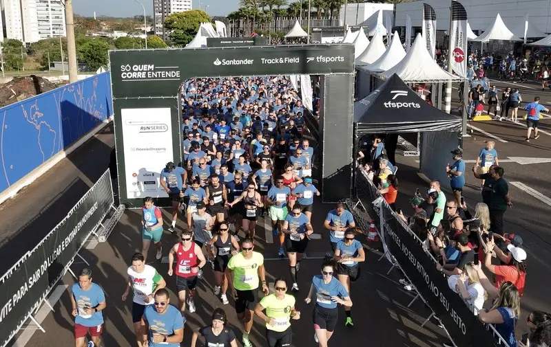largada de corrida com corredores passando por arco de largada com as marcas Santander e Track&Field, tendas e público ao lado