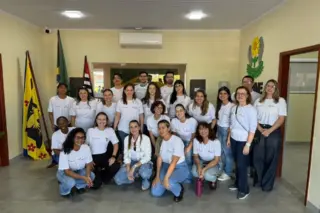 Grupo de voluntários posando em ambiente interno, usando camisetas brancas, com duas bandeiras ao fundo e um brasão na parede