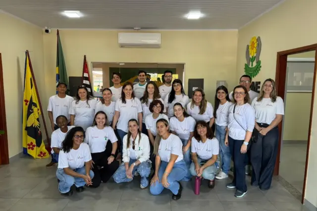 Grupo de voluntários posando em ambiente interno, usando camisetas brancas, com duas bandeiras ao fundo e um brasão na parede