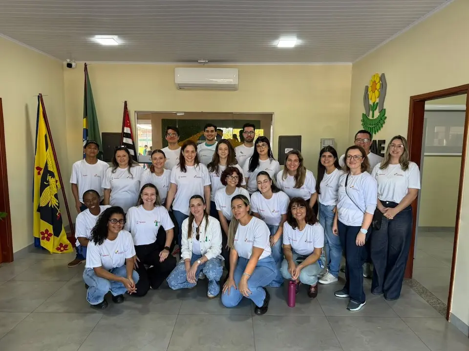 Grupo de voluntários posando em ambiente interno, usando camisetas brancas, com duas bandeiras ao fundo e um brasão na parede