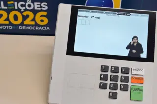 Urna eletrônica com visor mostrando tela 'Senador - 2ª vaga' e intérprete de libras; teclado numérico e botões 'BRANCO', 'CORRIGE' e 'CONFIRMA'; banner 'ELEIÇÕES 2026' ao fundo