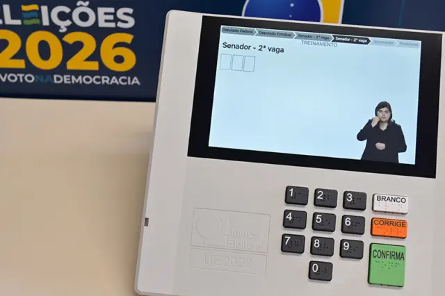 Urna eletrônica com visor mostrando tela 'Senador - 2ª vaga' e intérprete de libras; teclado numérico e botões 'BRANCO', 'CORRIGE' e 'CONFIRMA'; banner 'ELEIÇÕES 2026' ao fundo