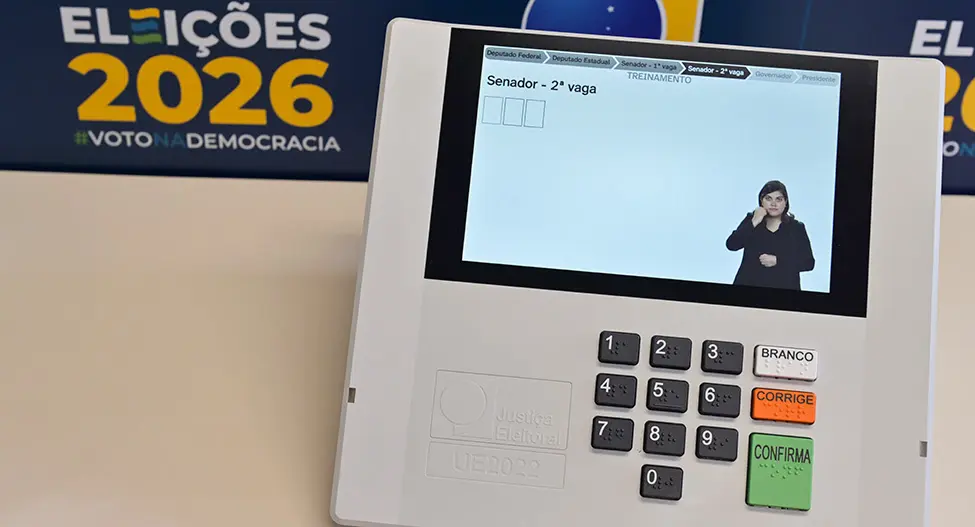 Urna eletrônica com visor mostrando tela 'Senador - 2ª vaga' e intérprete de libras; teclado numérico e botões 'BRANCO', 'CORRIGE' e 'CONFIRMA'; banner 'ELEIÇÕES 2026' ao fundo