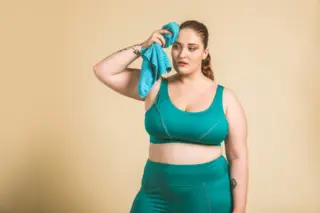 Mulher de corpo cheio vestindo top e legging verde-azulada, segurando uma toalha azul na testa contra fundo bege