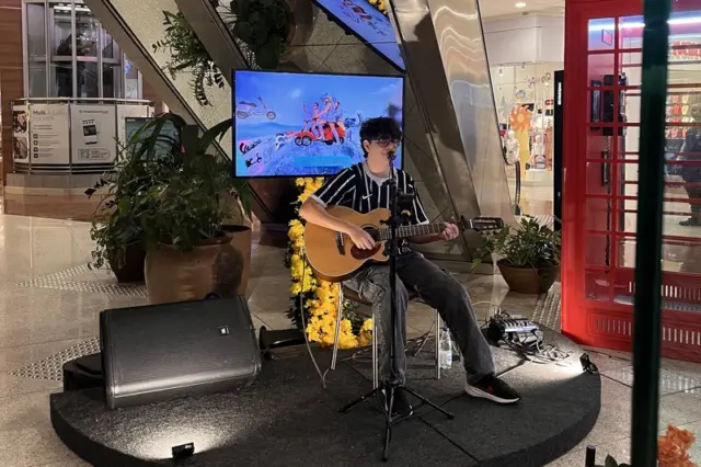 Artista sentado tocando violão em pequeno palco no piso térreo de um shopping, com microfone, monitor ao fundo e plantas