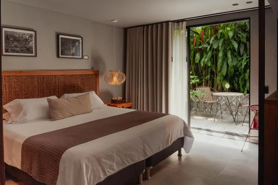 Quarto com cama de casal, luminária pendente e porta de correr aberta para área externa com plantas