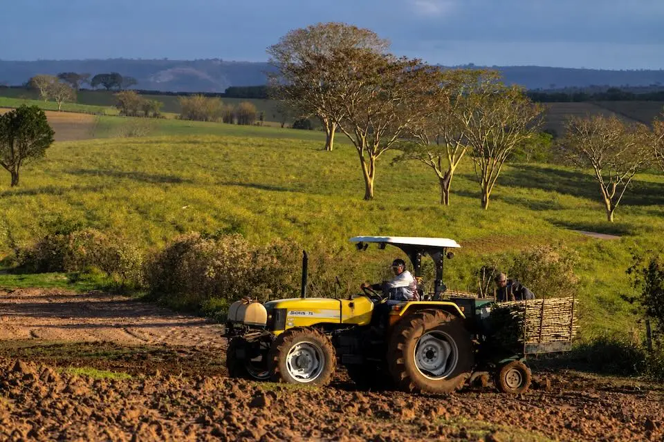 Trator agrícola amarelo em um campo arado com operador ao volante e implemento traseiro carregado, colinas verdes e árvores ao fundo