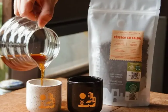 mão derramando café de uma jarra de vidro em duas xícaras sobre uma superfície de madeira; ao lado, um saco stand up pouch branco com janela mostrando grãos de café e rótulos com QR codes