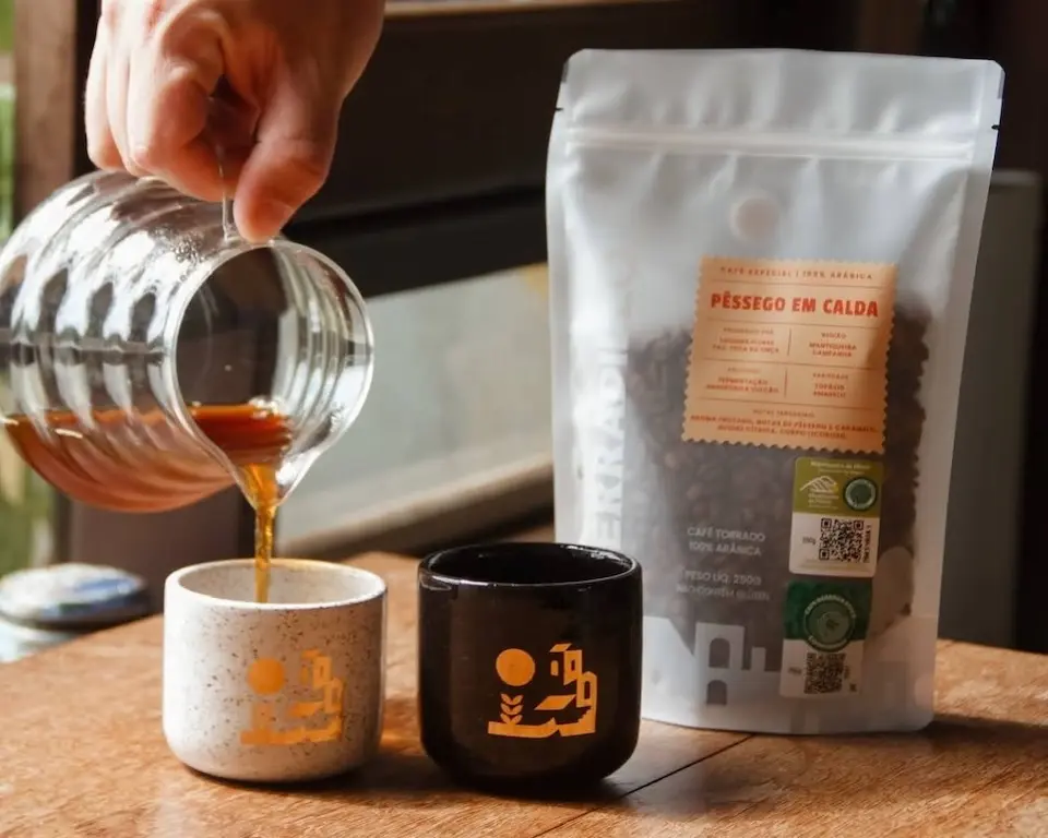 mão derramando café de uma jarra de vidro em duas xícaras sobre uma superfície de madeira; ao lado, um saco stand up pouch branco com janela mostrando grãos de café e rótulos com QR codes