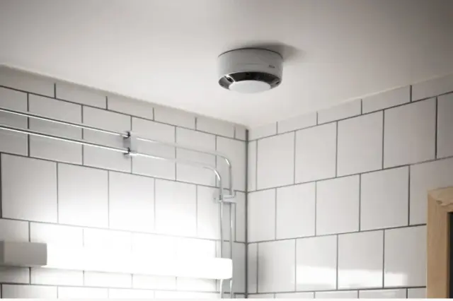 Sensor circular branco montado no teto de um ambiente com azulejos brancos, luminária de parede e eletrodutos metálicos