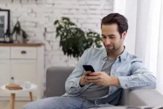 Homem adulto sentado em um sofá olhando para um smartphone que segura com as duas mãos