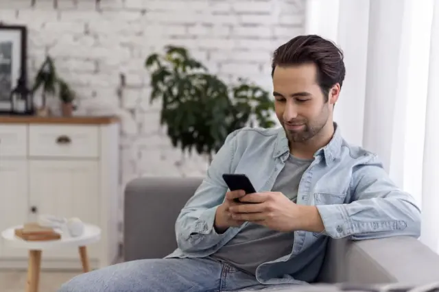 Homem adulto sentado em um sofá olhando para um smartphone que segura com as duas mãos