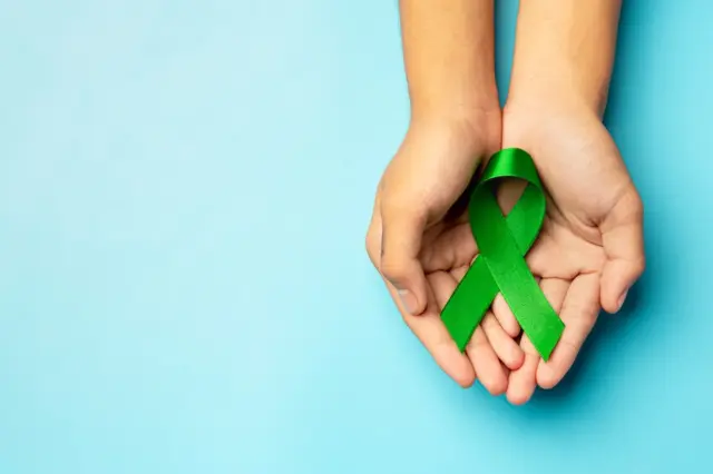 Duas mãos abertas segurando um laço verde de conscientização sobre um fundo azul claro