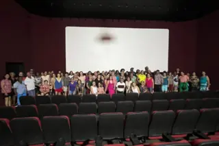 Grupo de pessoas, em maioria idosas, em pé em frente a uma tela branca de cinema; fileiras de poltronas vazias em primeiro plano