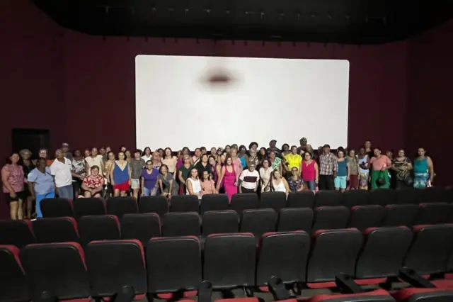 Grupo de pessoas, em maioria idosas, em pé em frente a uma tela branca de cinema; fileiras de poltronas vazias em primeiro plano