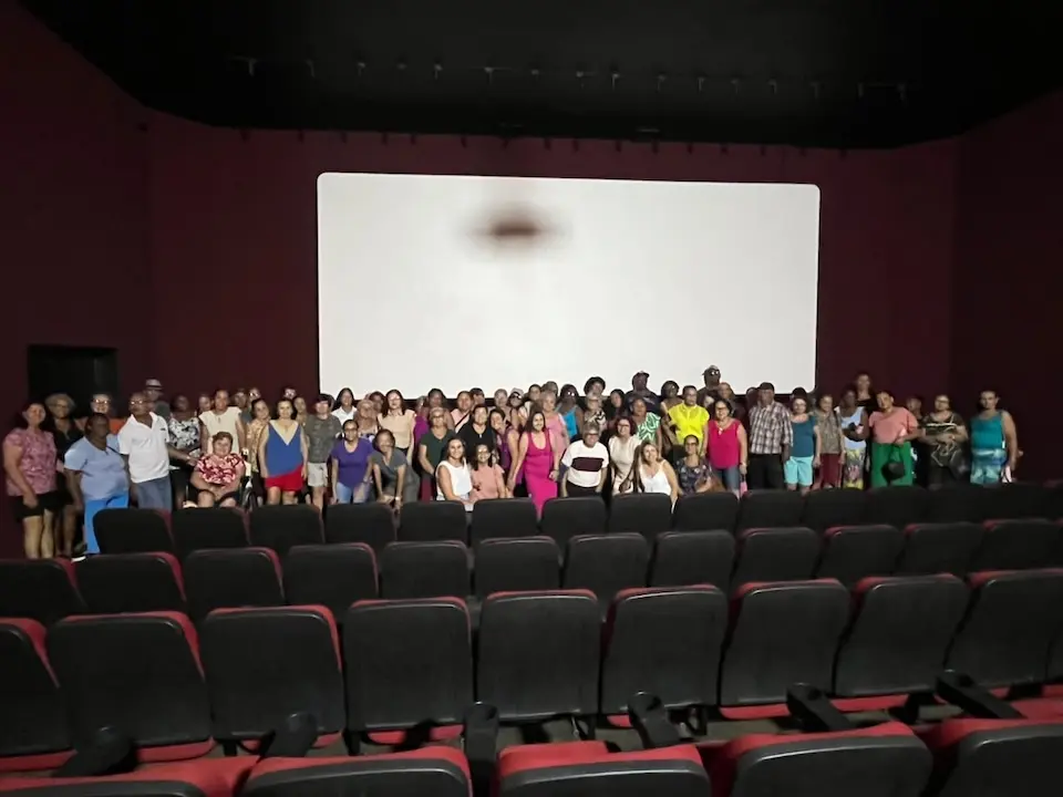 Grupo de pessoas, em maioria idosas, em pé em frente a uma tela branca de cinema; fileiras de poltronas vazias em primeiro plano