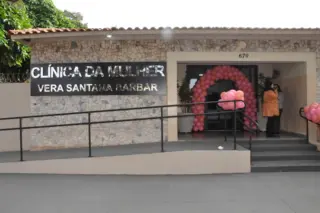 Fachada da Clínica da Mulher Vera Santana Barbar com placa metálica, rampa de acesso e arco de balões rosa na entrada; duas pessoas próximas à porta