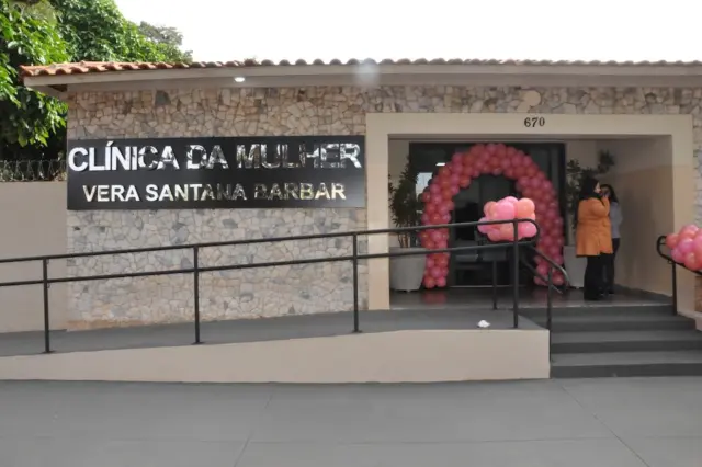 Fachada da Clínica da Mulher Vera Santana Barbar com placa metálica, rampa de acesso e arco de balões rosa na entrada; duas pessoas próximas à porta