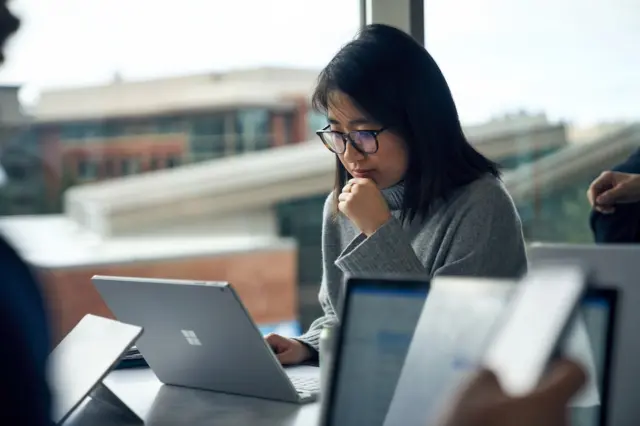 Mulher jovem com óculos usando um laptop Microsoft Surface em uma mesa, com outros dispositivos e janela com prédio desfocado ao fundo